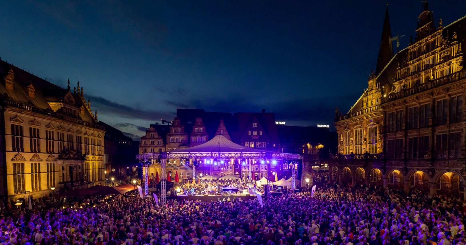 Open Air auf dem Marktplatz mit großer, runder, überdachter Bühne auf dem ein Orchester spielt